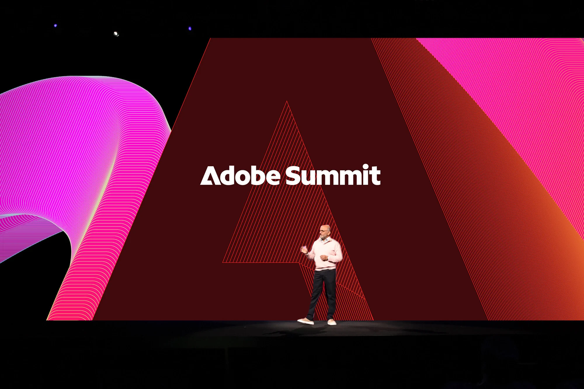 Adult male presenter on stage at the Adobe Summit, with a branded backdrop featuring a linear pink geometric pattern on black.