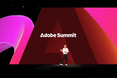 Speaker on a large dark stage presenting in front of a vibrant pink and red geometric graphic screen with the Adobe Summit logo