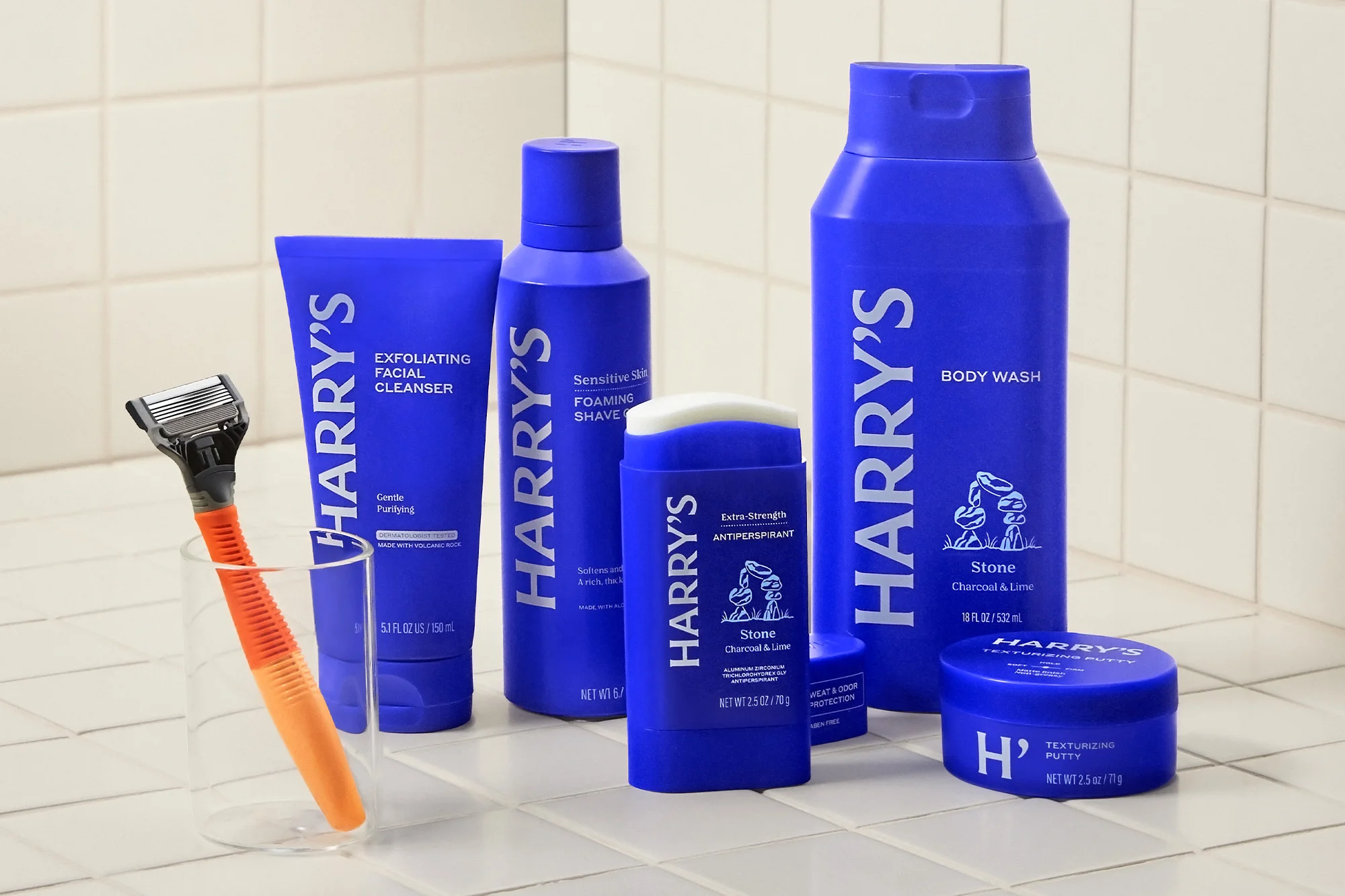 Still life shot of Harry's brand men's grooming products in blue packaging on a tile surface, including shave, cleanser, and body wash.