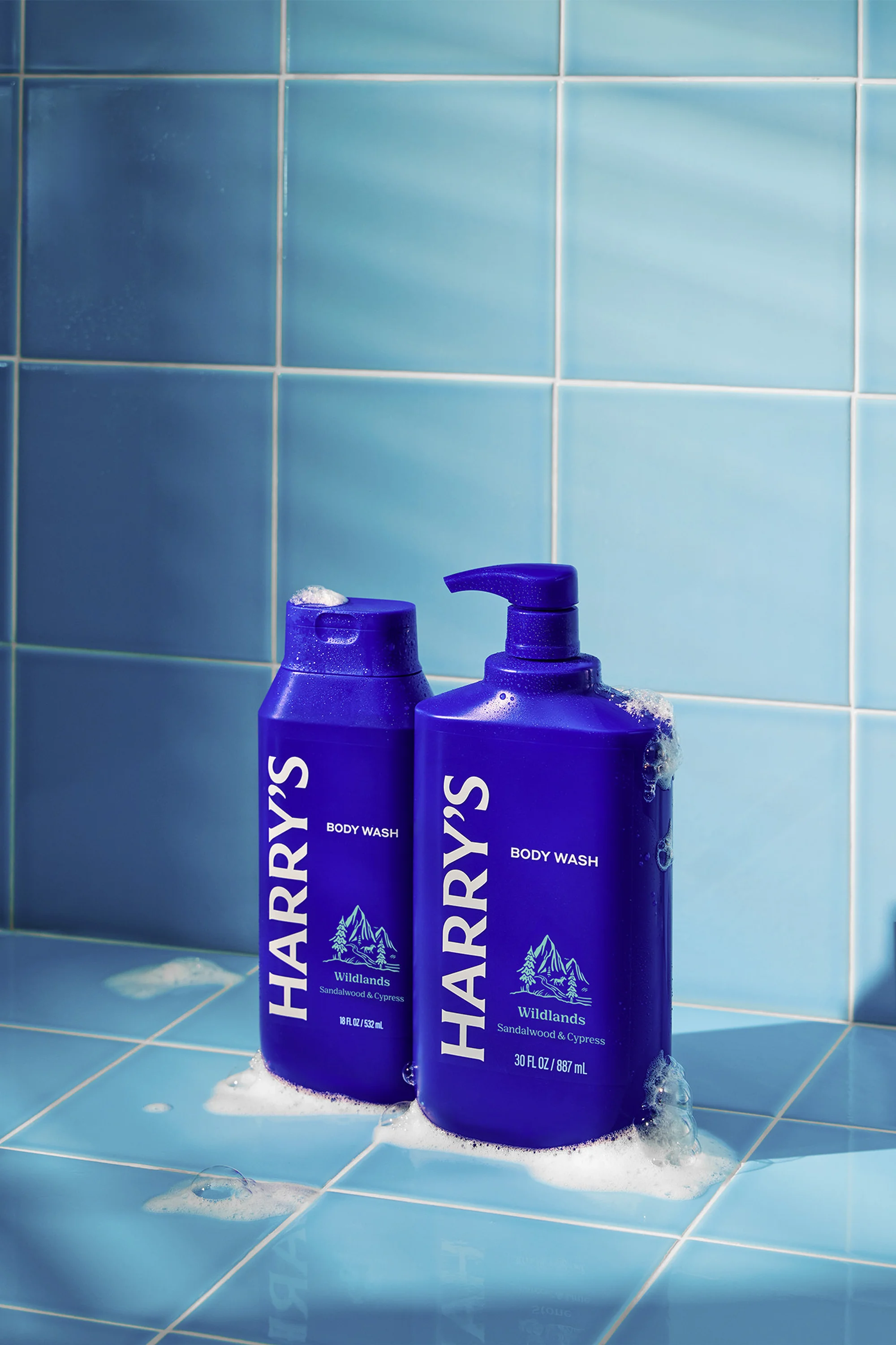 Two blue Harry's brand body wash bottles sit on a light blue tile surface with soap bubbles. The taller bottle has a pump dispenser.