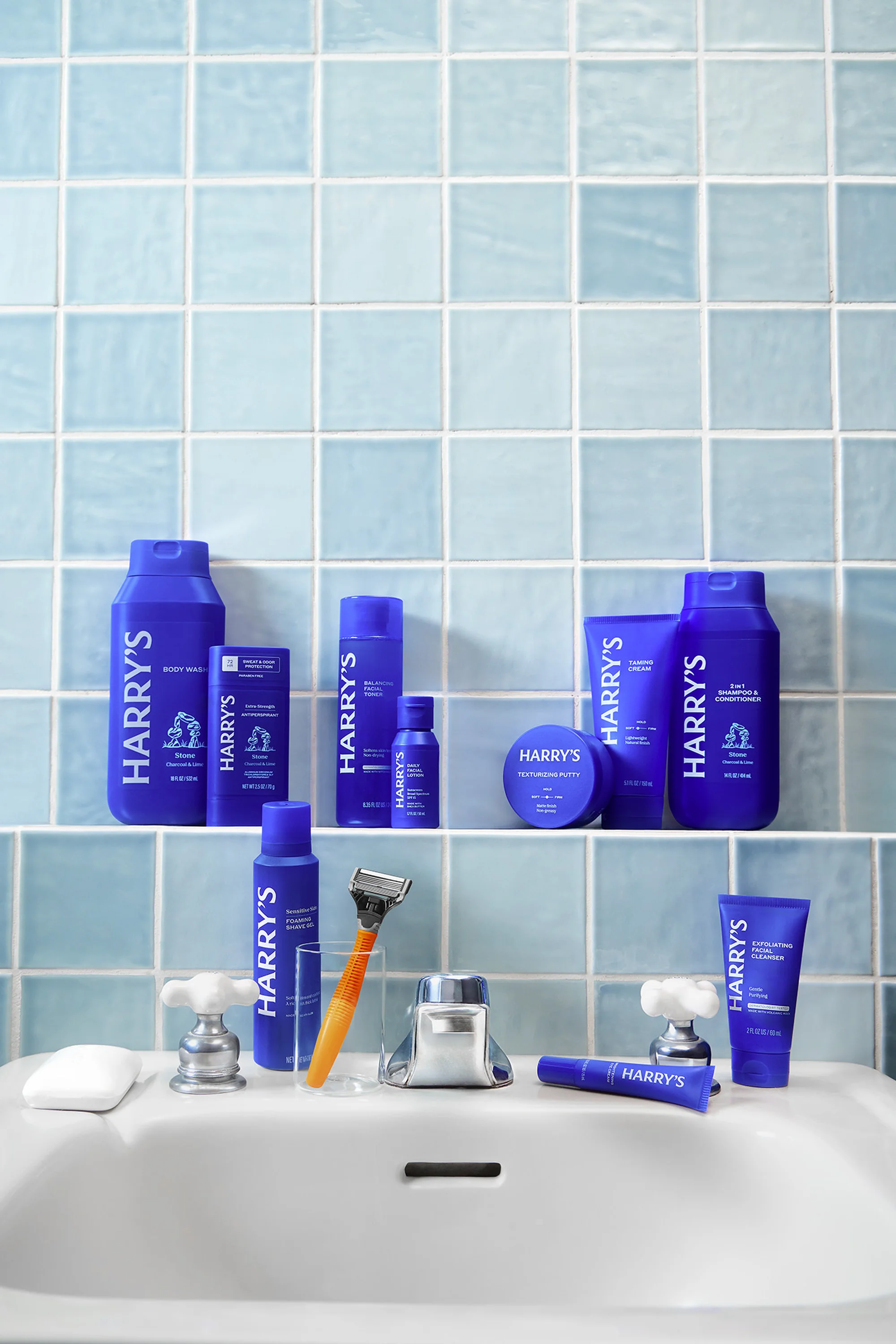 A collection of Harry's brand grooming products are neatly arranged on a white bathroom sink with a light blue tiled backsplash.