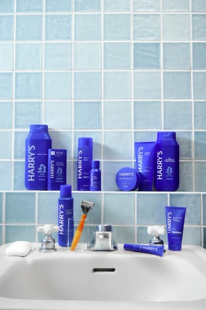 A collection of Harry's brand grooming products are neatly arranged on a white bathroom sink with a light blue tiled backsplash.