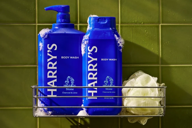 Two blue bottles of Harry's brand body wash are on a wire rack, with bubbly foam on the bottles against a tiled green wall.