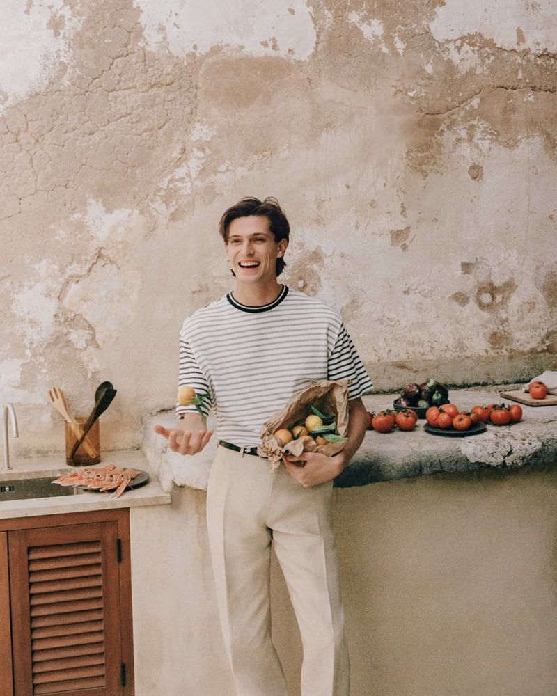 A young adult man wearing a striped t-shirt and light pants stands in a kitchen, holding a bag of lemons and juggling a lemon.