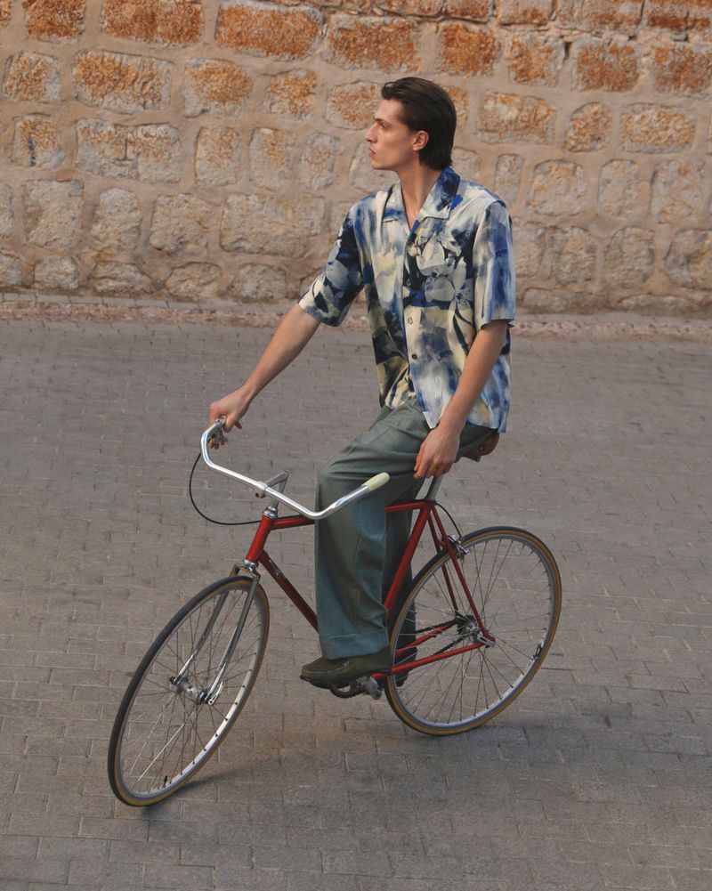 A young adult male rides a red vintage road bicycle on a brick-paved street with a stone wall in the background during daytime.