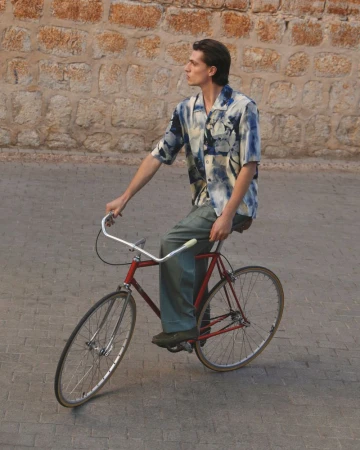 A young adult male rides a red vintage road bicycle on a brick-paved street with a stone wall in the background during daytime.