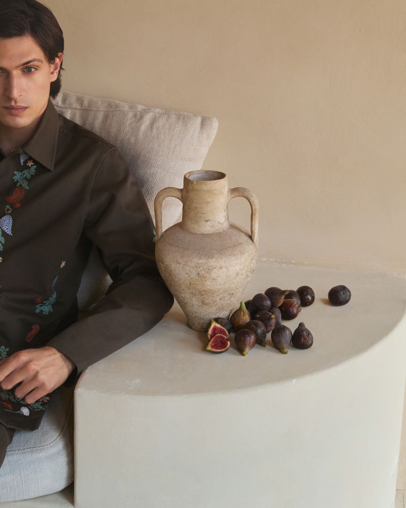 Man in olive shirt poses next to antique vase and figs on a light beige table against matching wall in an indoor setting.