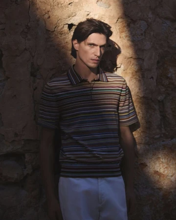 Man in a striped shirt posing near a stone wall, with dramatic lighting enhancing the textures and shadows.