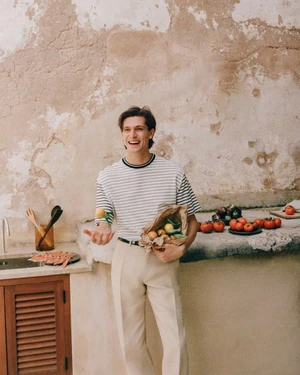 Smiling man wearing a striped t-shirt and light trousers holding a bag of produce in a rustic kitchen setting.