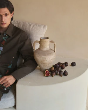 Model wearing embroidered olive green shirt next to a ceramic amphora and scattered figs on a white surface
