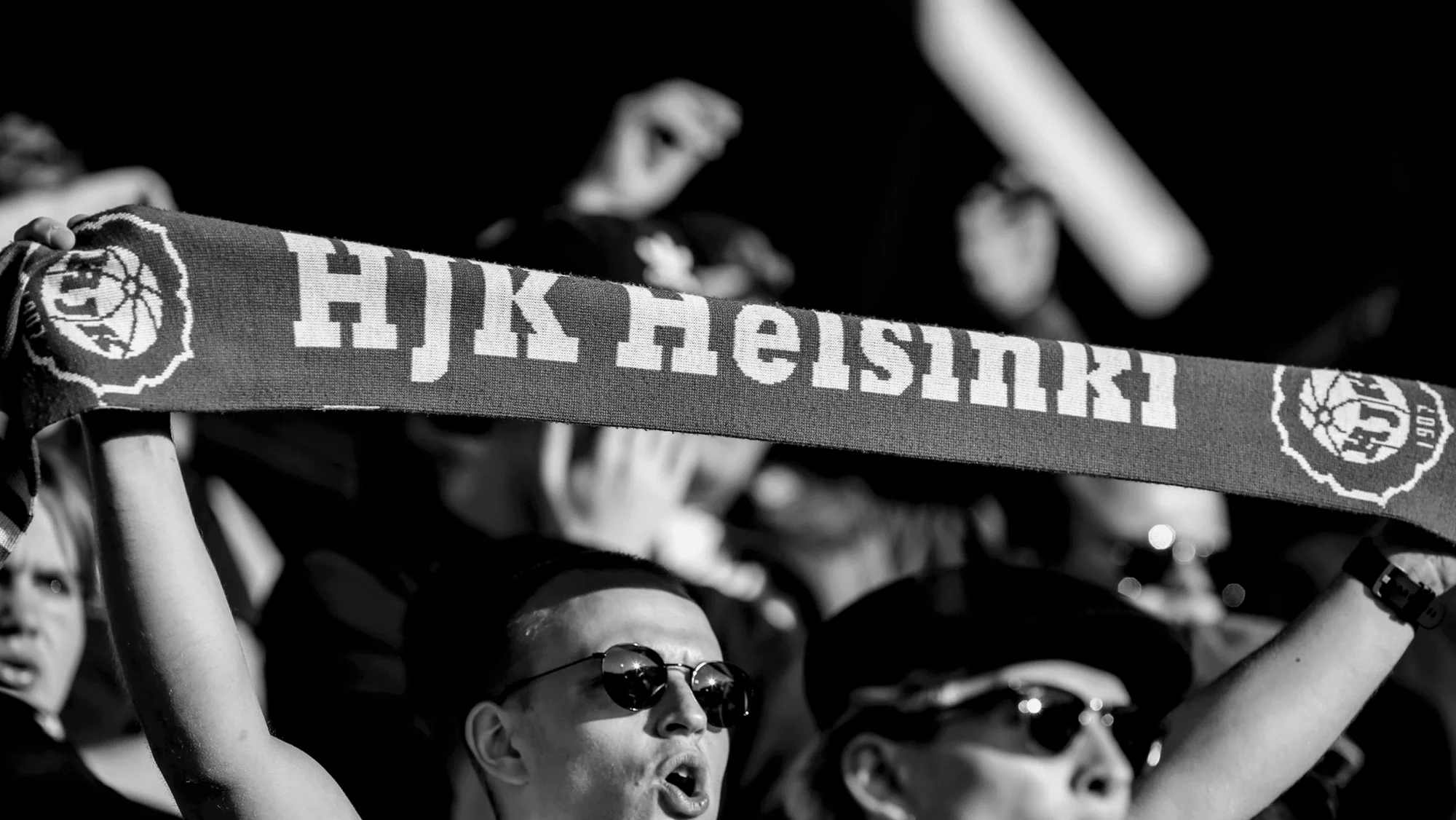 Black and white photo of sports fans holding a scarf with "HJK Helsinki" text, showing support and excitement in an indoor setting.