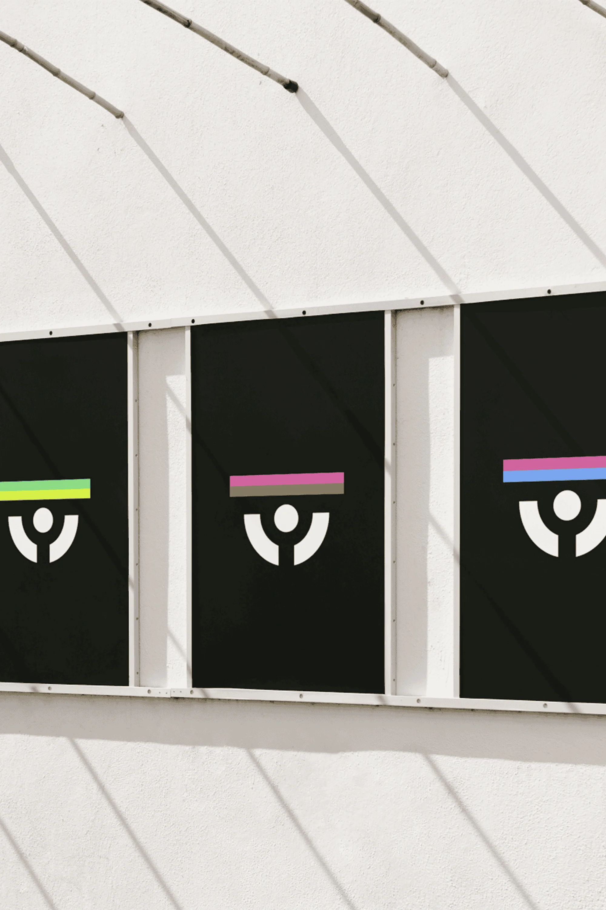 Three black display panels featuring white graphic logos and coloured horizontal bars mounted on a white wall under harsh sunlight.