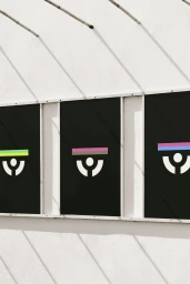 Three black display panels featuring white graphic logos and coloured horizontal bars mounted on a white wall under harsh sunlight.