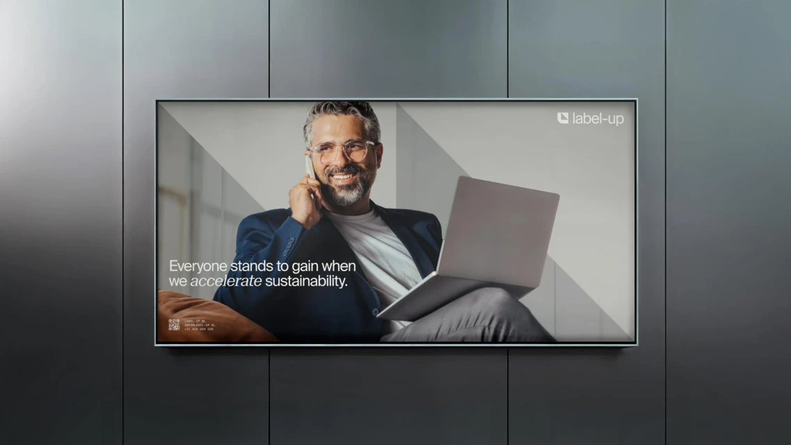 Professional man smiles on a phone call, holding a laptop, in an ad promoting sustainability, with gray wall background.