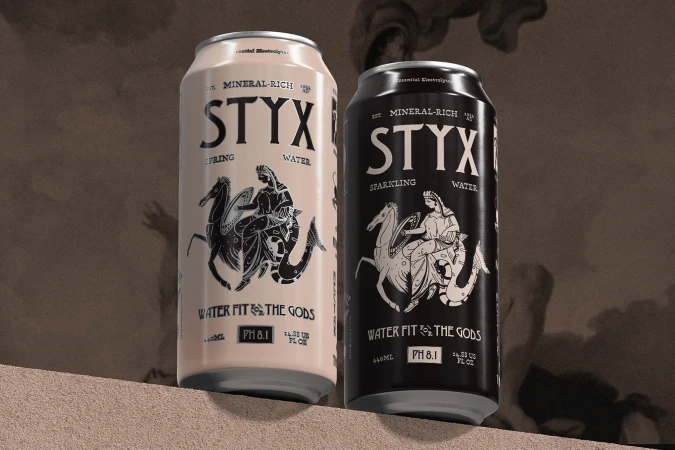 Two cans of STYX brand spring water, one beige and one black, sitting on a tan surface against a blurred background.