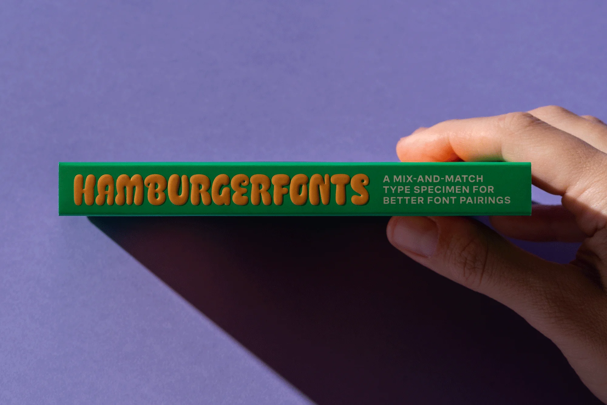 Hand holds a green box with the word 'HAMBURGERFONTS' printed in orange on it. The background is a solid lilac color.