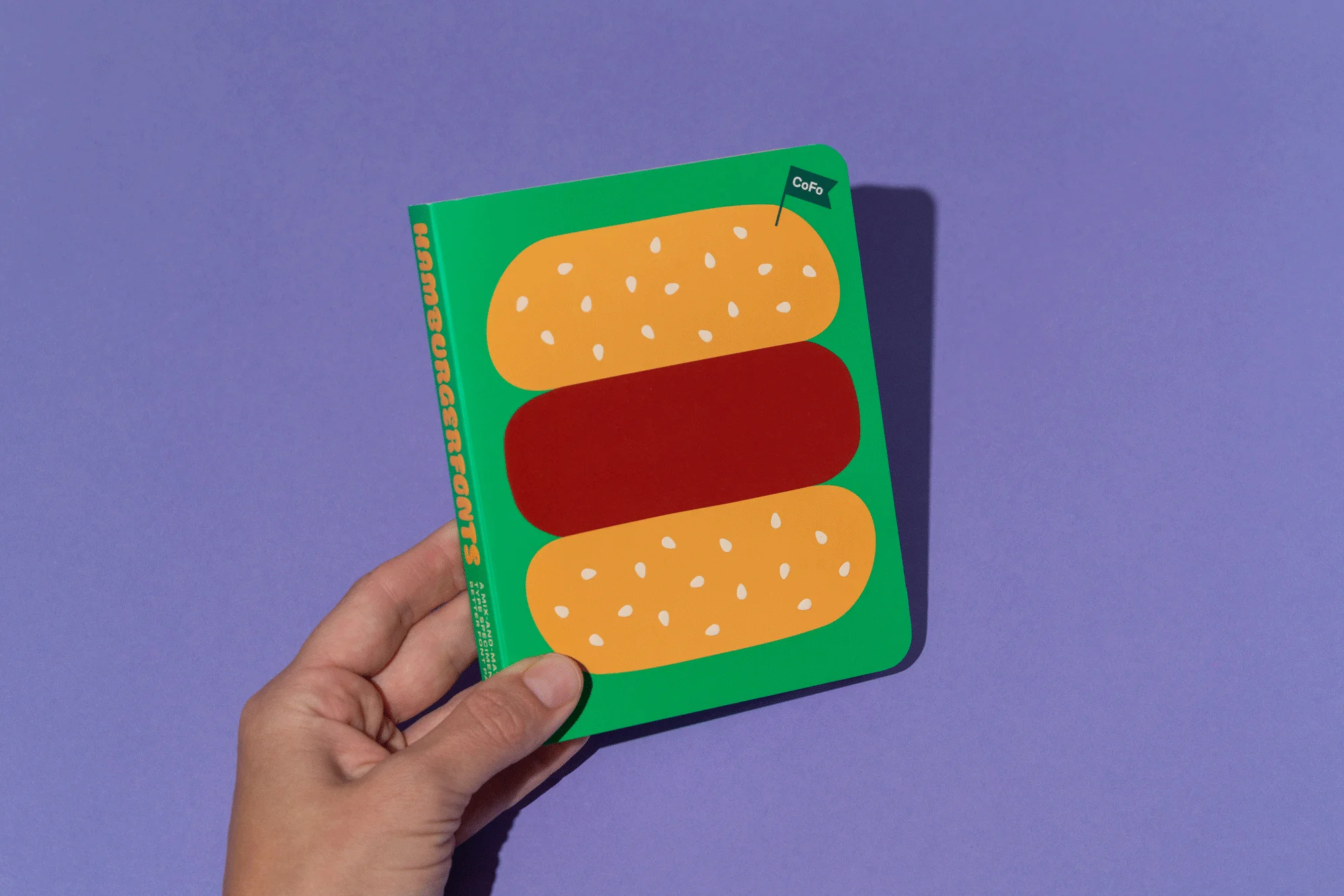 A hand holds a green notebook with a burger illustration against a lilac background. The notebook has a CoFo logo in the corner.
