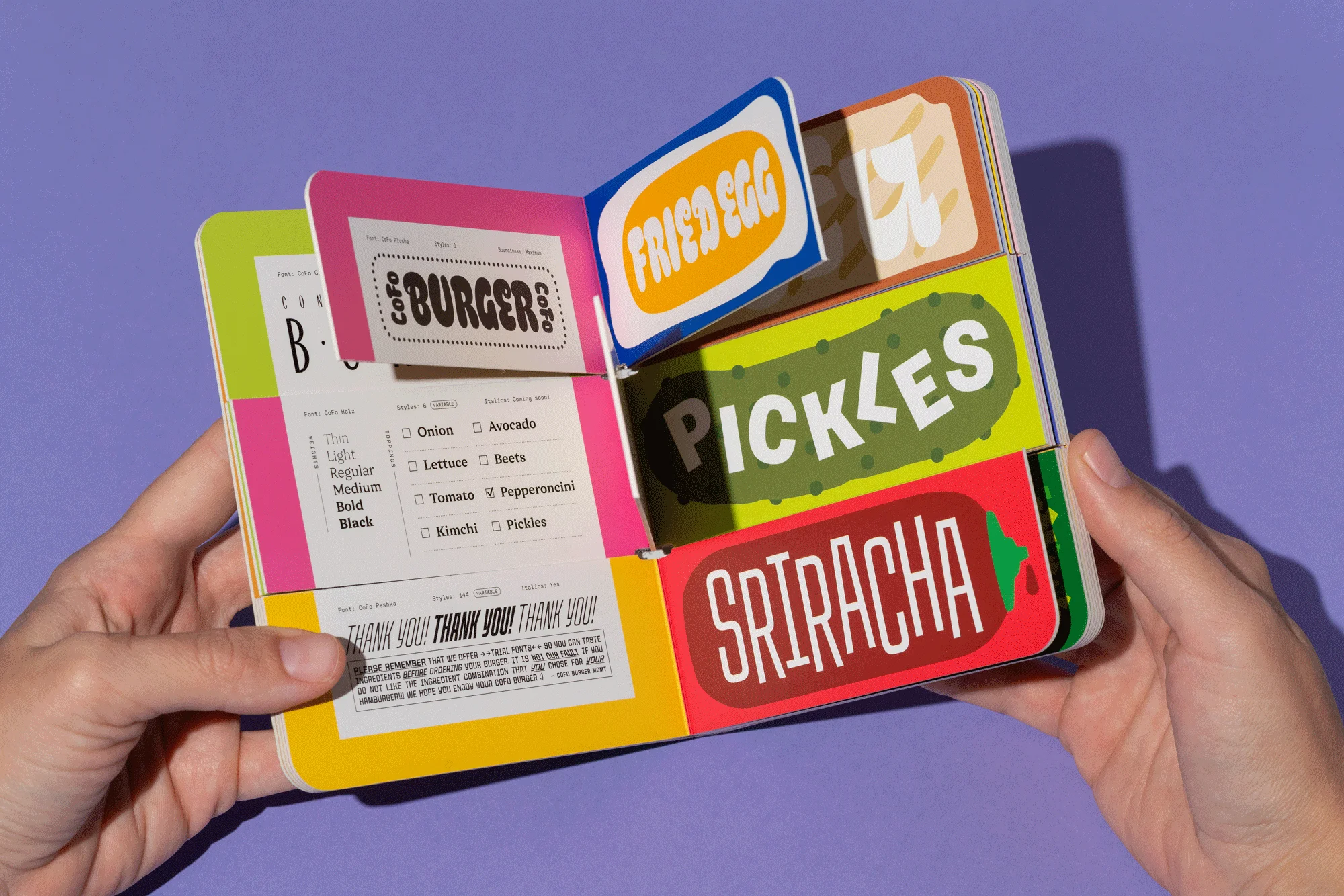 Hands hold an open colorful board book titled 'Burger' with illustrated burger ingredients like fried egg, pickles, and sriracha.