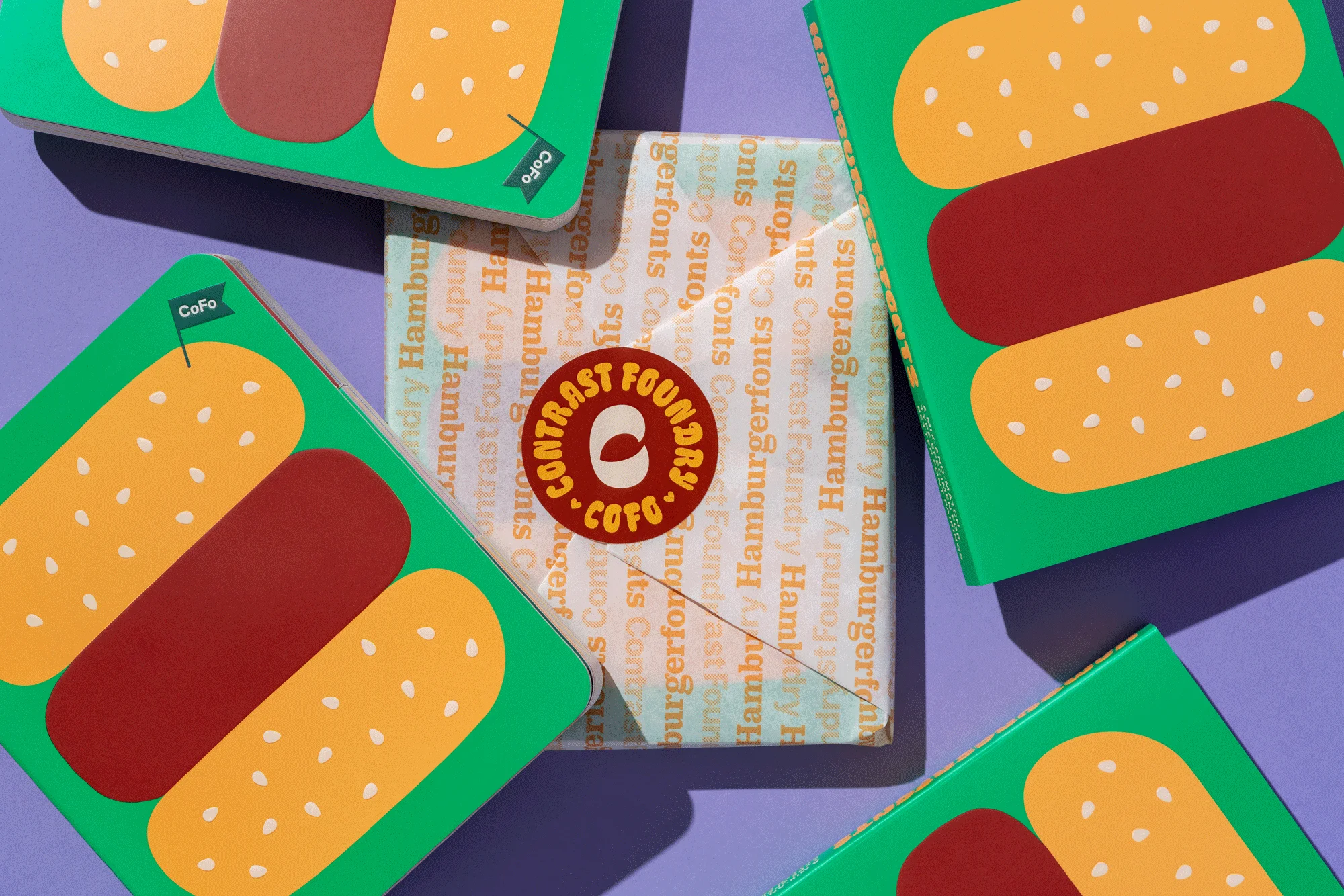 Overhead studio shot of promotional product packaging featuring a hamburger graphic and the Contrast Foundry logo on a lavender background.