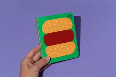 Hand holding a small green notebook with a hamburger illustration against a solid purple background.
