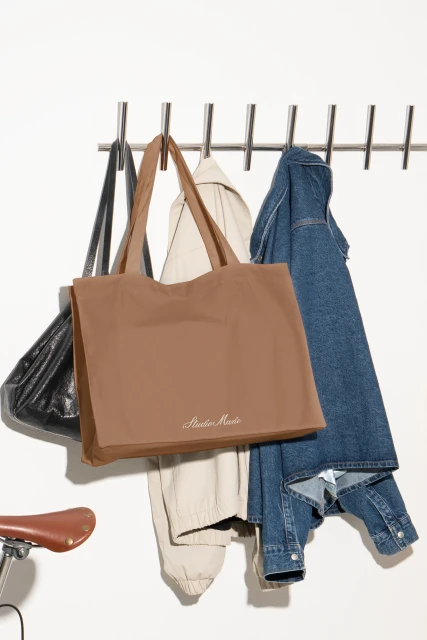 Brown tote bag hanging on a metal rack with a black purse, beige pants, and a blue denim jacket against a white background.
