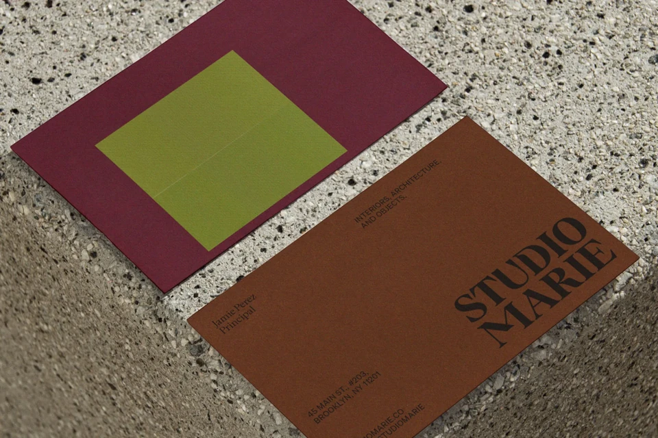 Two business cards, one in a plum color with a green square, and one in brown with black text, rest on a textured surface.