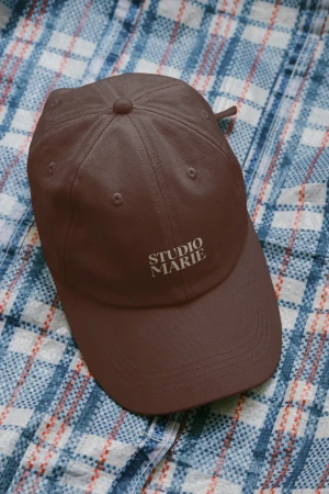 Brown baseball cap with 'Studio Marie' text on a blue and white plaid surface, showcasing casual headwear and studio branding.