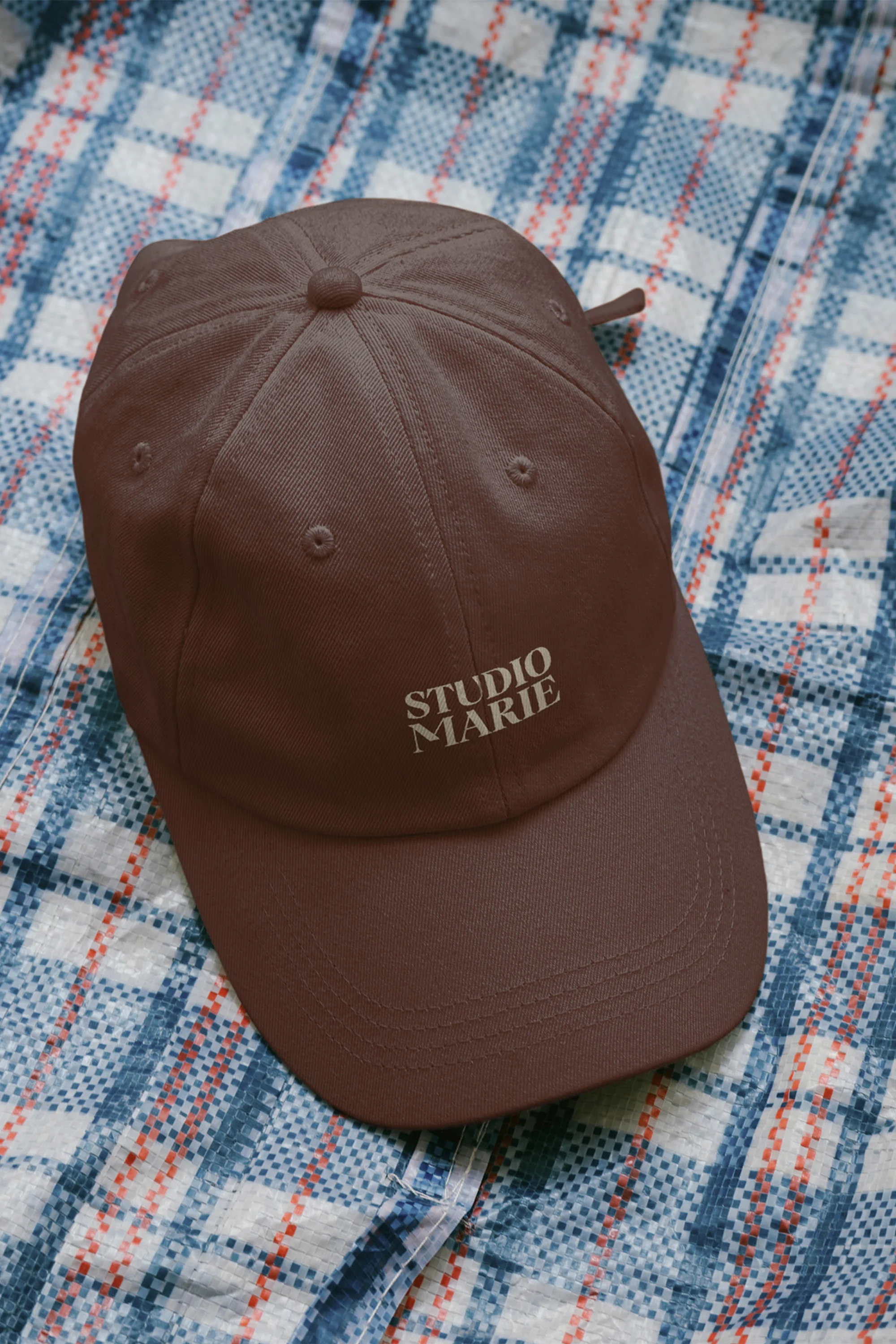 Brown baseball cap featuring the Studio Marie wordmark resting on a blue plaid shirt background.