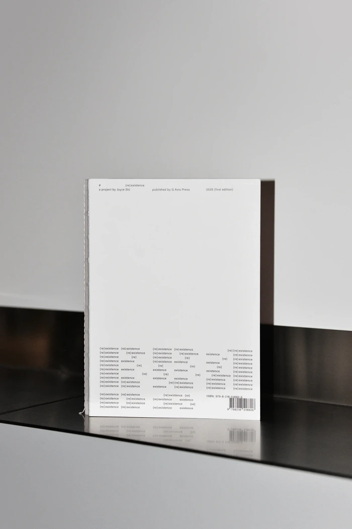 A minimalist white softcover book standing upright on a sleek dark metallic shelf against a neutral grey wall.