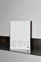 A minimalist white softcover book standing upright on a sleek dark metallic shelf against a neutral grey wall.