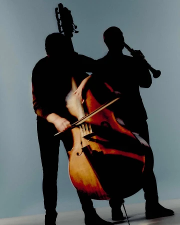 Two musicians are seen in silhouette playing a cello and a clarinet in front of a light blue background, bathed in soft, diffused lighting.