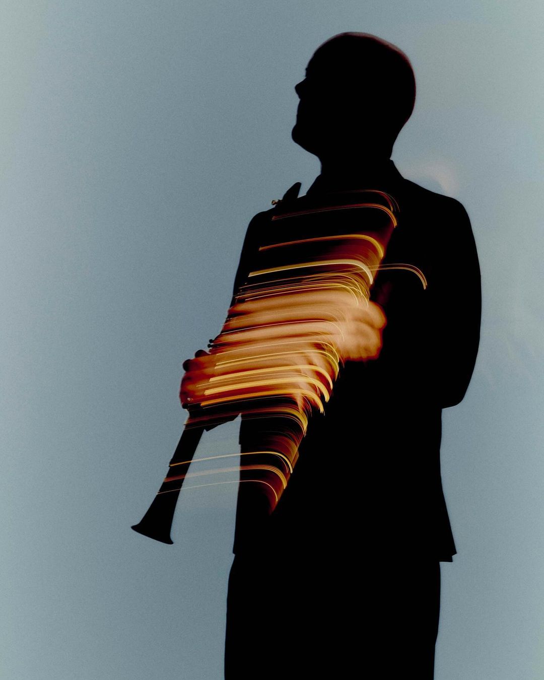 Silhouette of a bald man playing a clarinet with light streaks, creating a sense of motion and musical energy against a grey background.