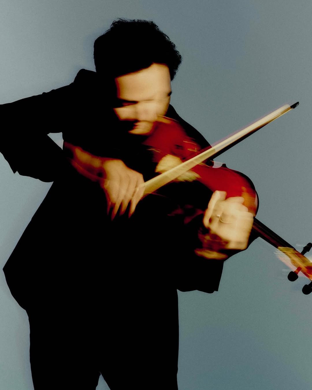 An adult male is seen playing a violin in a long-exposure studio shot, with a focus on the violin and the motion of the bow.
