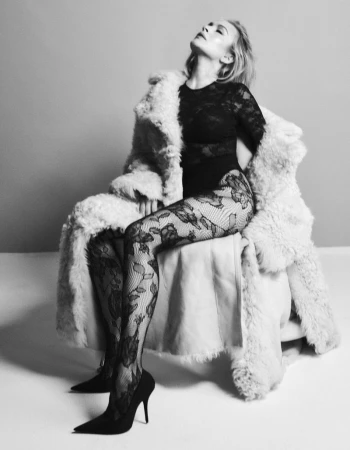 Black and white shot of a woman in a fur coat, lace bodysuit, and floral tights sitting on a stool, posing in a photo studio.