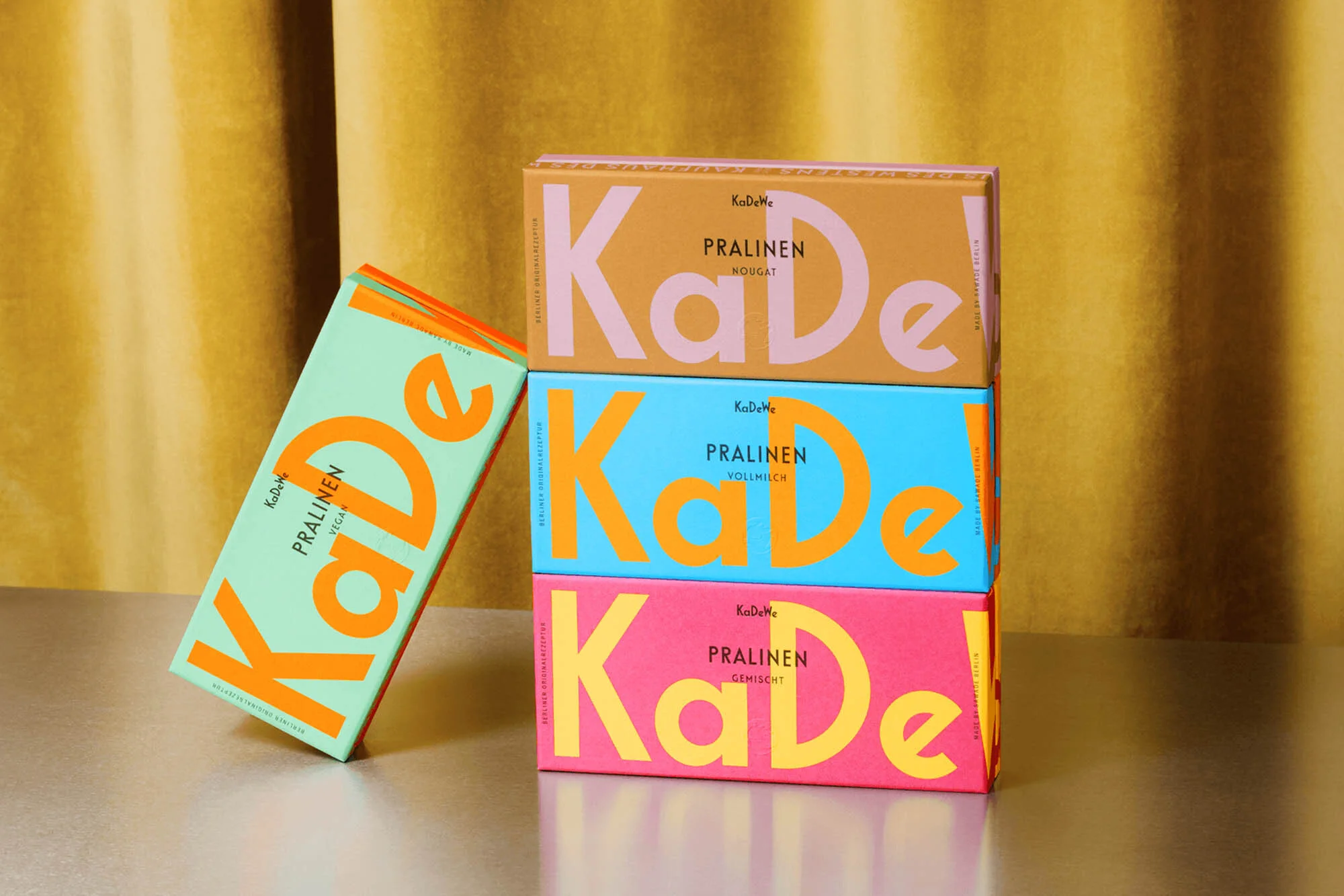 Three colorful boxes of KaDe pralines are stacked and leaning against a mustard-yellow curtain backdrop, creating a vibrant and tempting display.