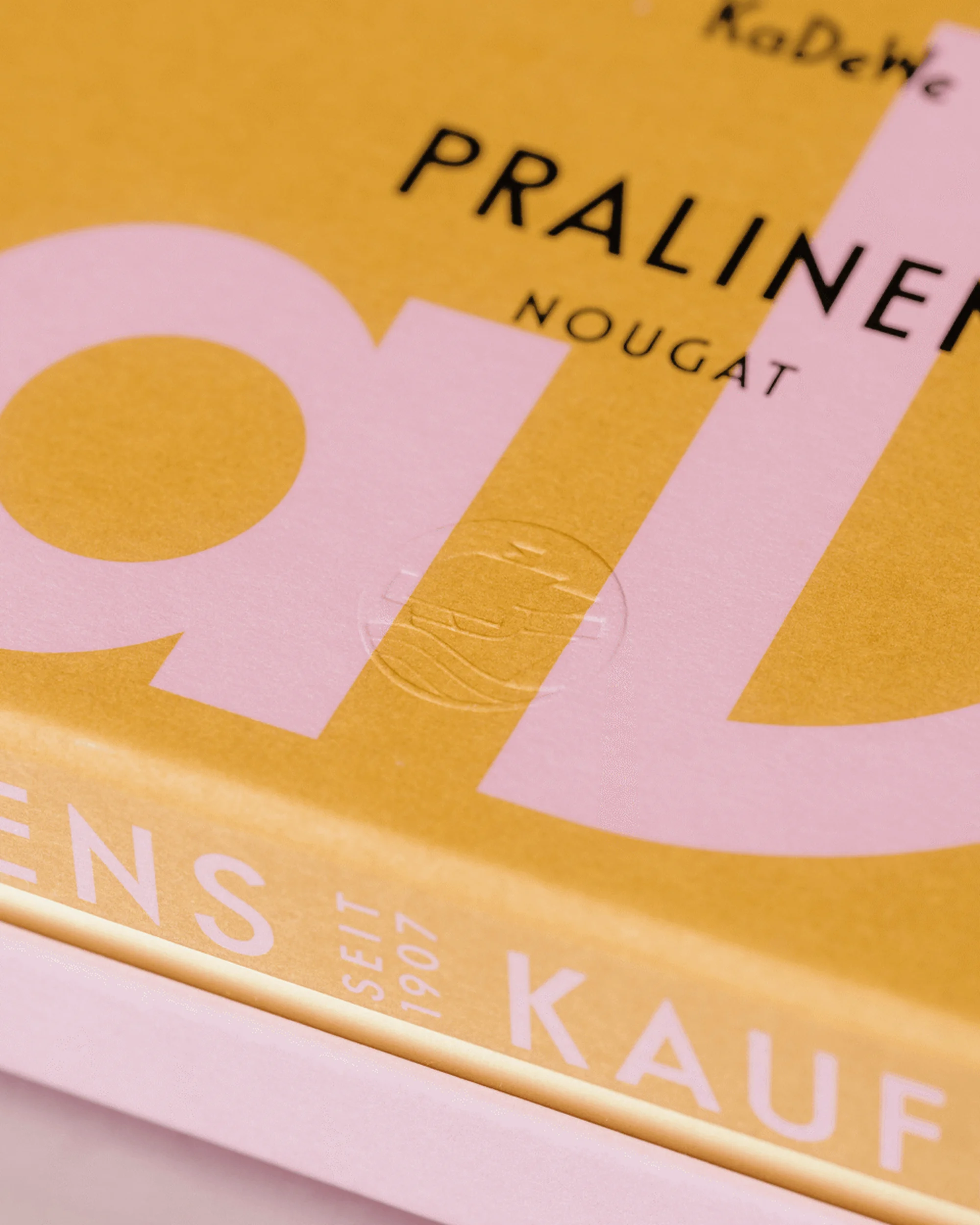 Close-up of a box of Pralinen Nougat from KaDeWe, featuring pink and yellow colors, emphasizing the brand and product name.