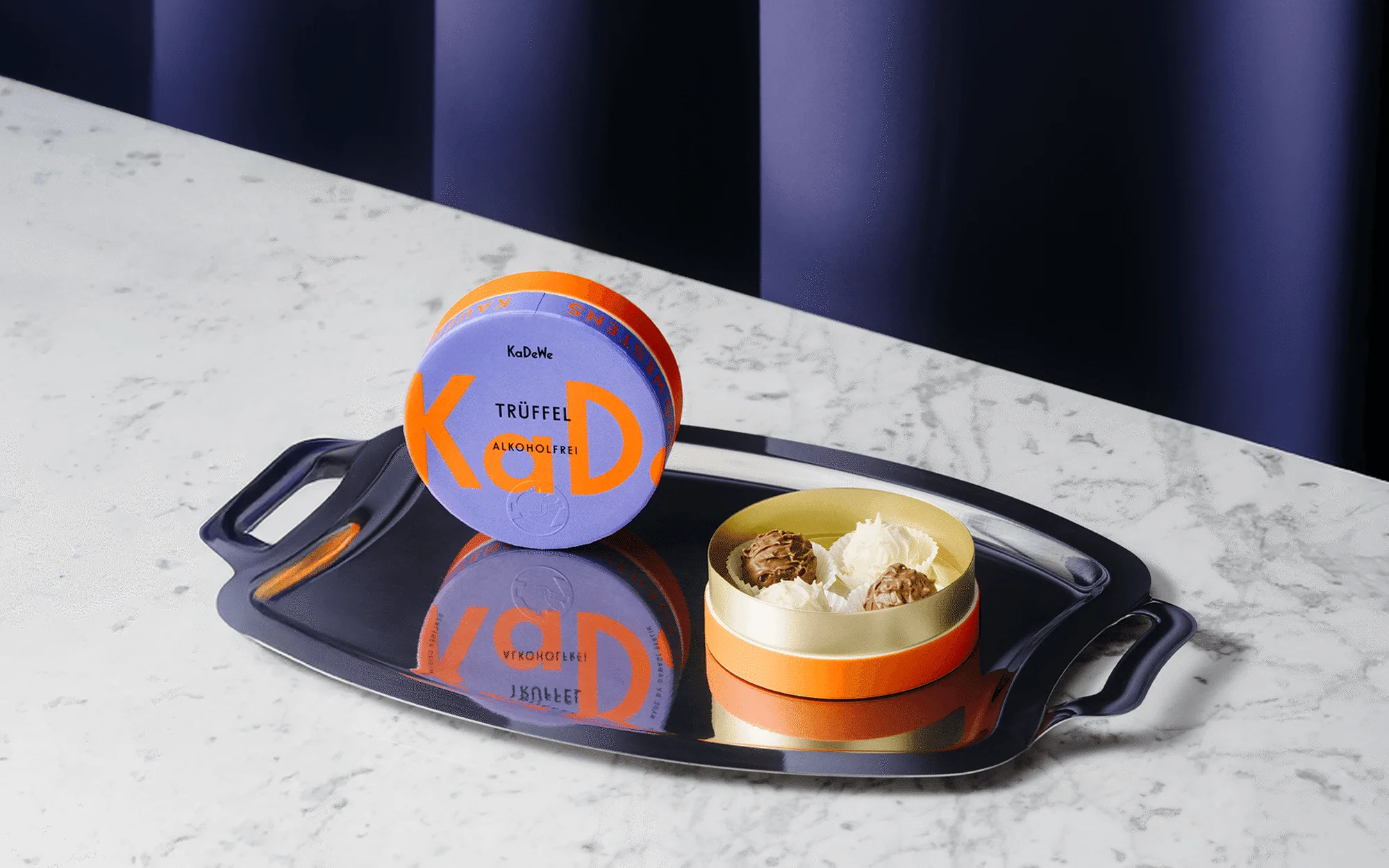 Studio shot of a purple truffle box with orange text, accompanied by a round box filled with chocolate truffles on a silver tray.