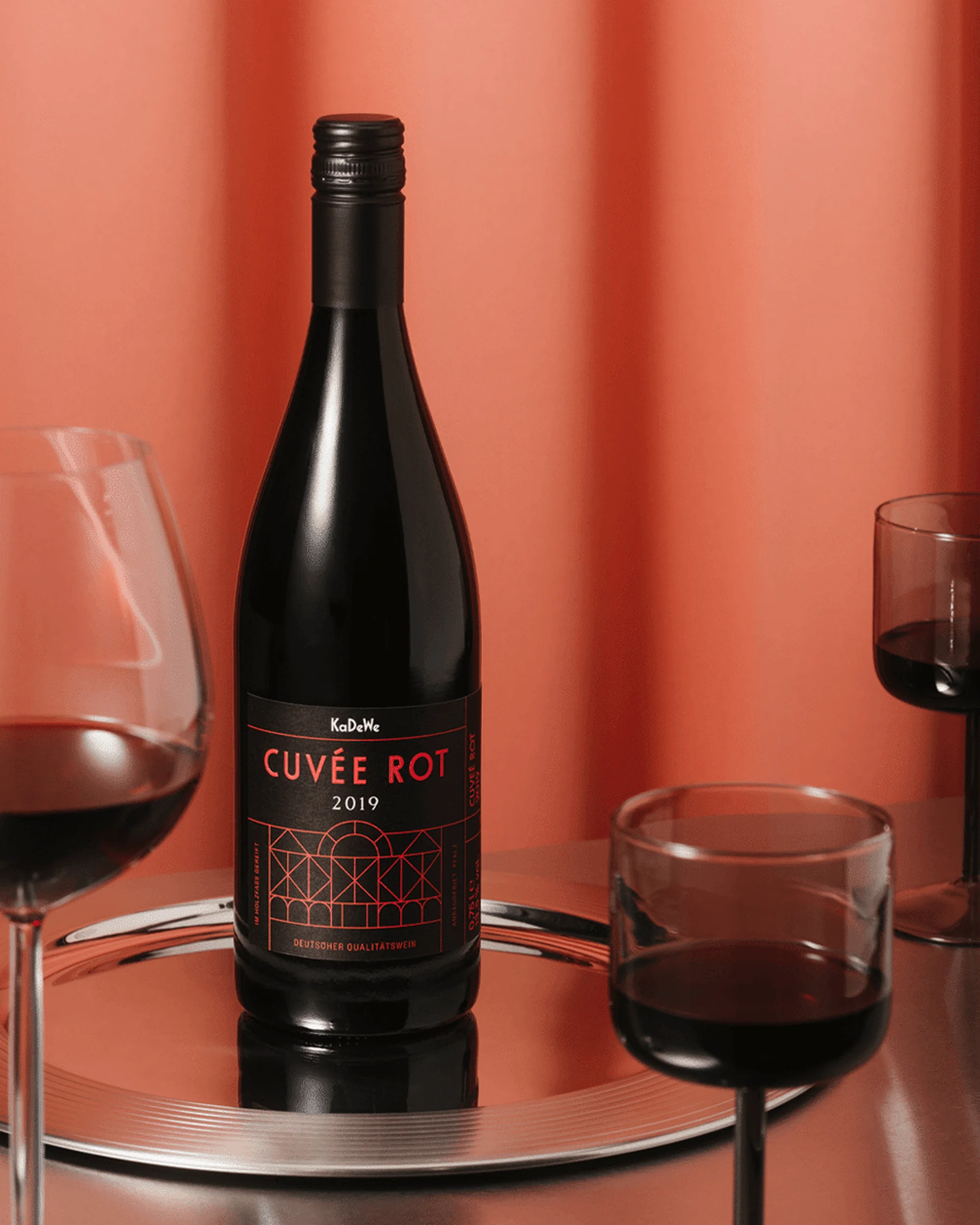 A dark wine bottle labeled Cuvée Rot standing on a reflective silver tray, surrounded by three glasses filled with red wine.