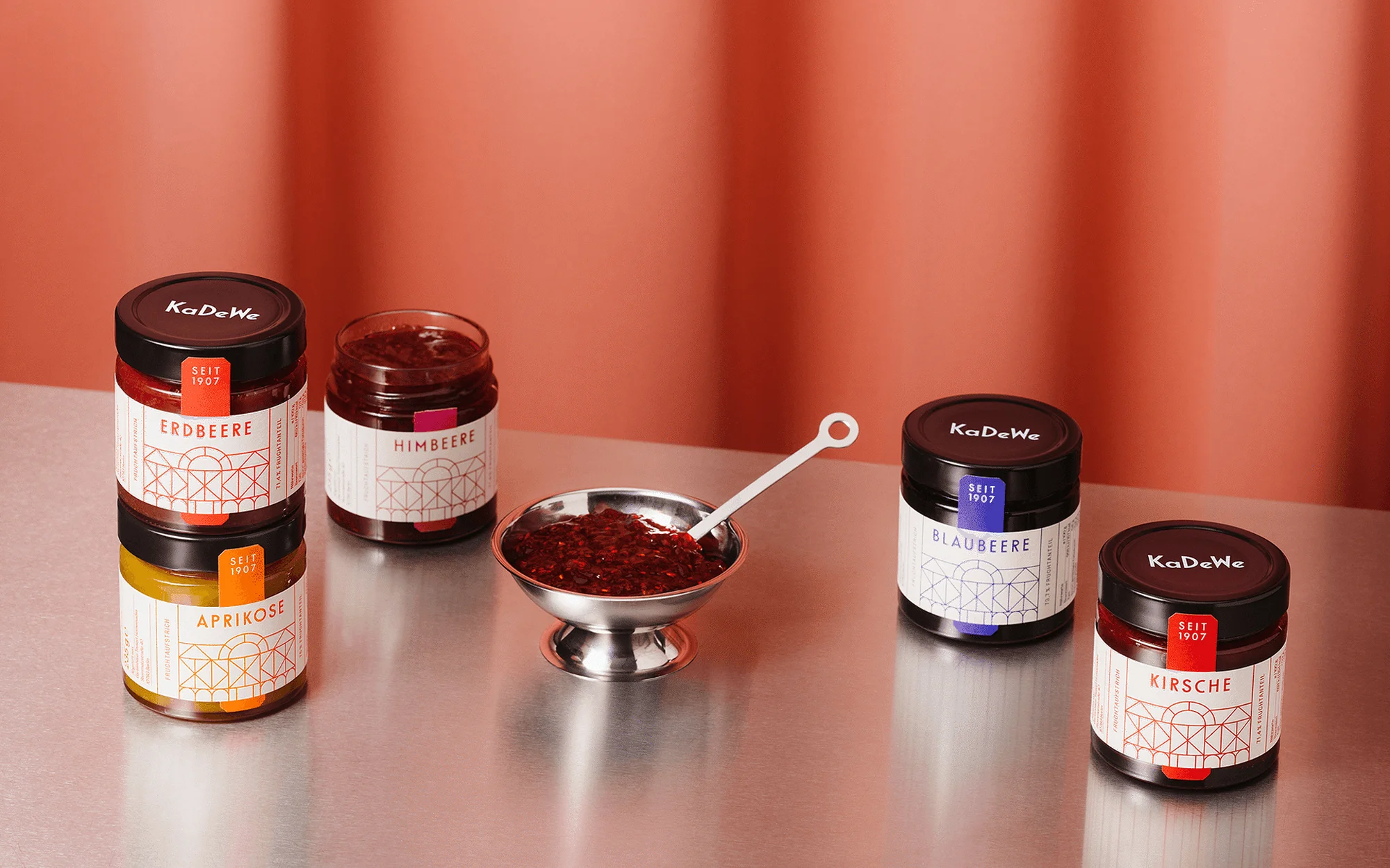 Arrangement of KaDeWe brand fruit preserves in jars and a silver bowl with spoon against an orange backdrop in a studio setting.