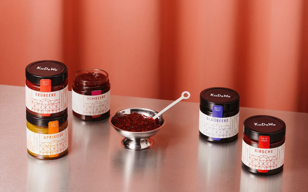Arrangement of KaDeWe brand fruit preserves in jars and a silver bowl with spoon against an orange backdrop in a studio setting.