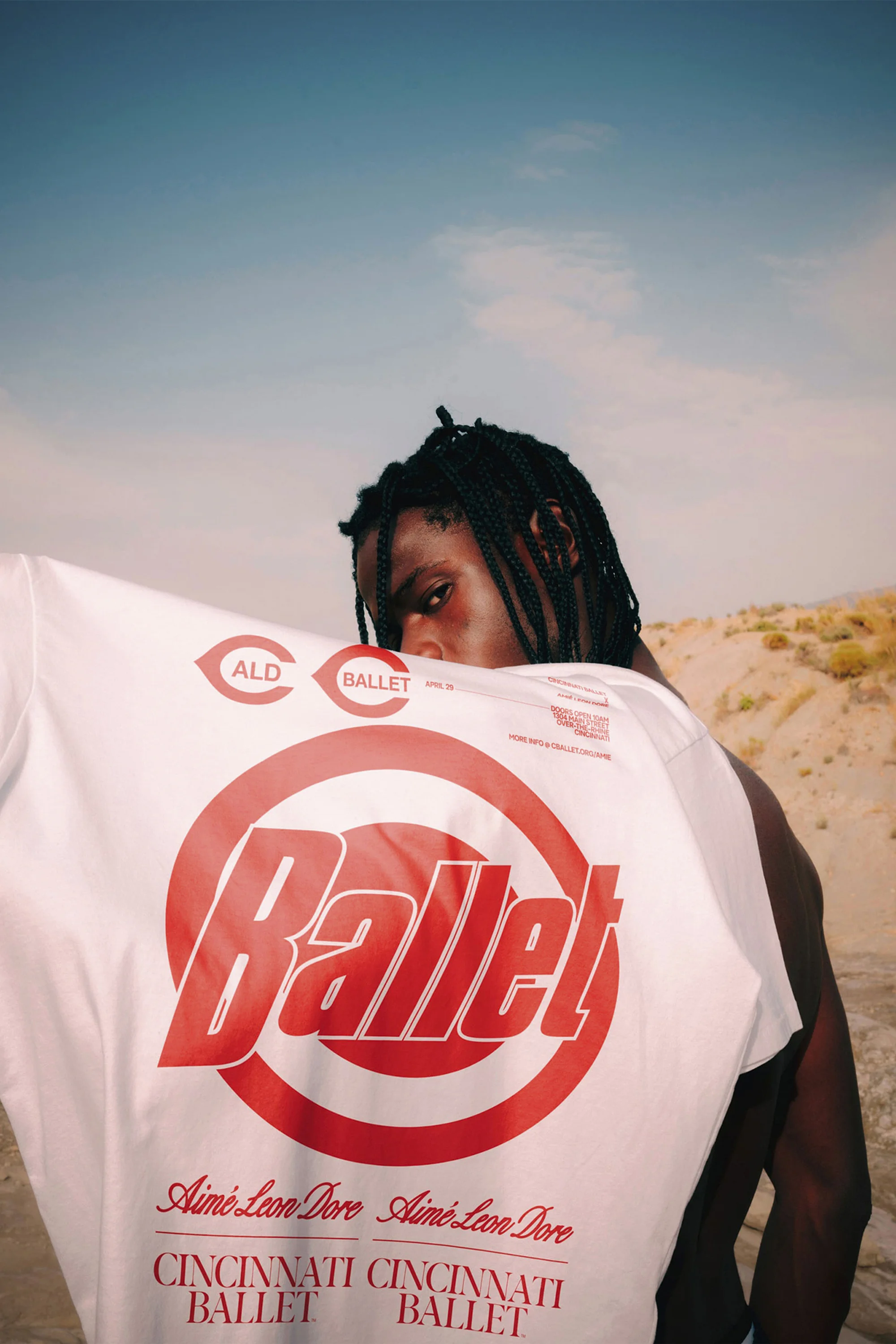 An adult male poses in an outdoor, desert-like environment while holding a white t-shirt with a bold 'Ballet' design in red.