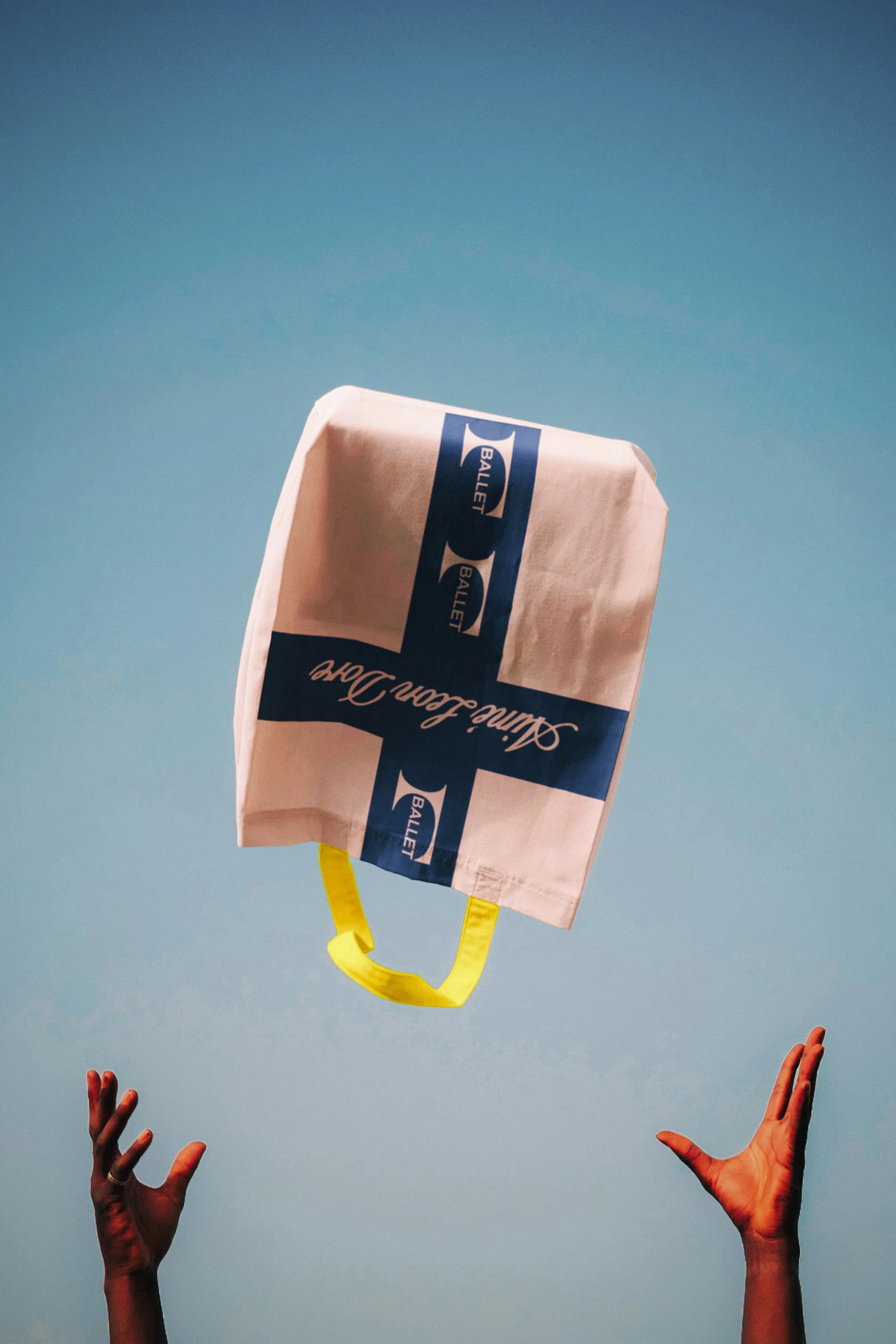 Tote bag with blue branding tossed in the air against a clear blue sky, framed by outstretched hands below.