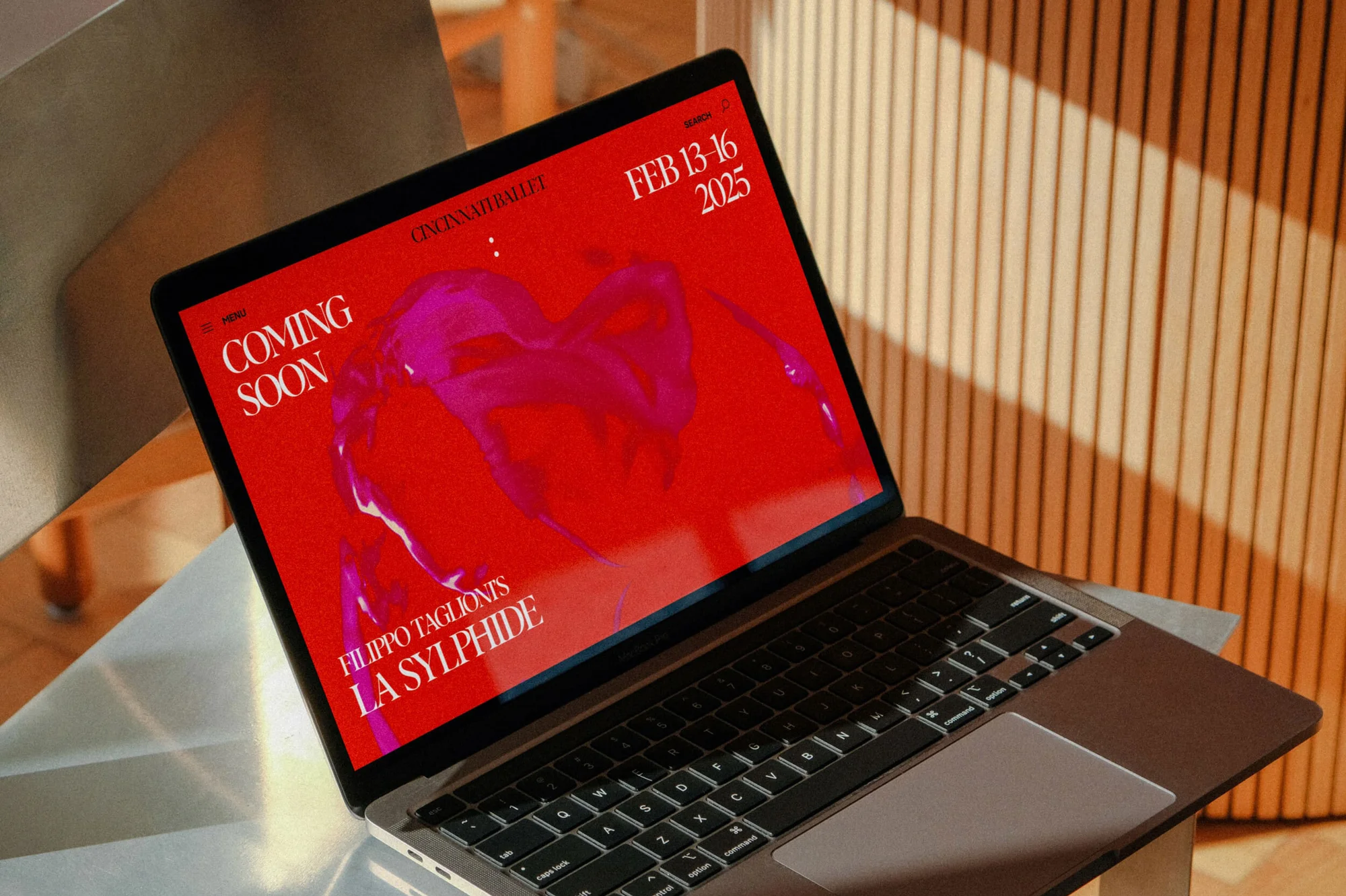 Laptop displaying Cincinnati Ballet's 'Coming Soon' page featuring a red, abstract image with event dates Feb 13-16, 2025, next to a wooden wall.