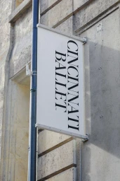 Vertical sign reading Cincinnati Ballet mounted on a stone building exterior next to a dark pole