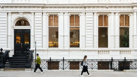 Exterior facade of an ornate white neoclassical building with black double doors, tall windows, and pedestrians walking past on the sidewalk.