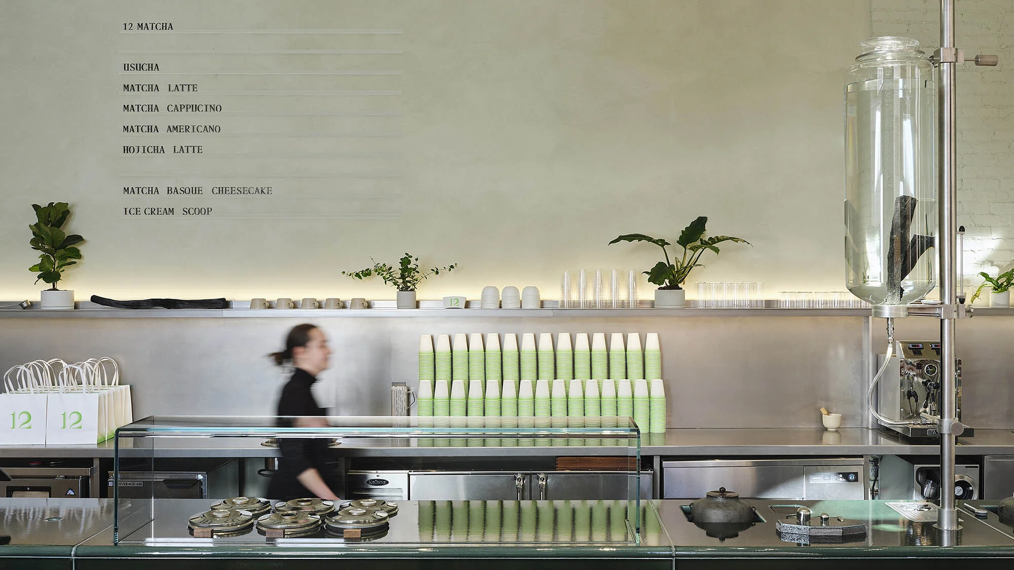 Interior view of a modern cafe with a server walking past a counter filled with green paper cups and beverage-making equipment.