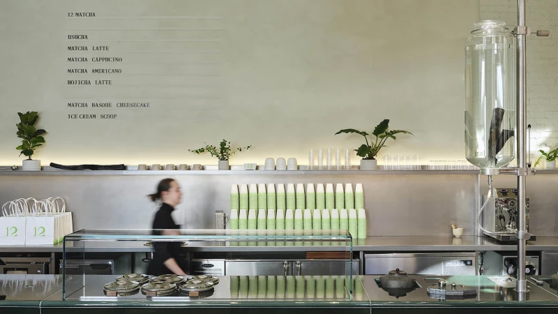 Interior view of a modern cafe with a server walking past a counter filled with green paper cups and beverage-making equipment.