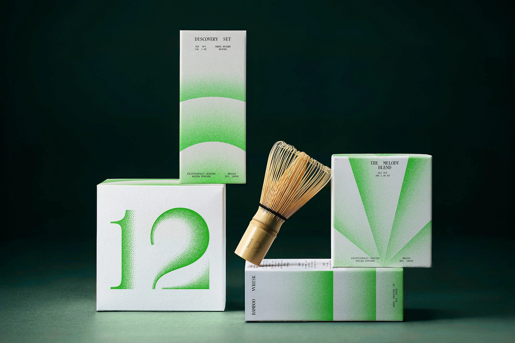 Close-up studio shot of a matcha whisk next to product boxes with green and white branding and minimalist design against a dark green backdrop.
