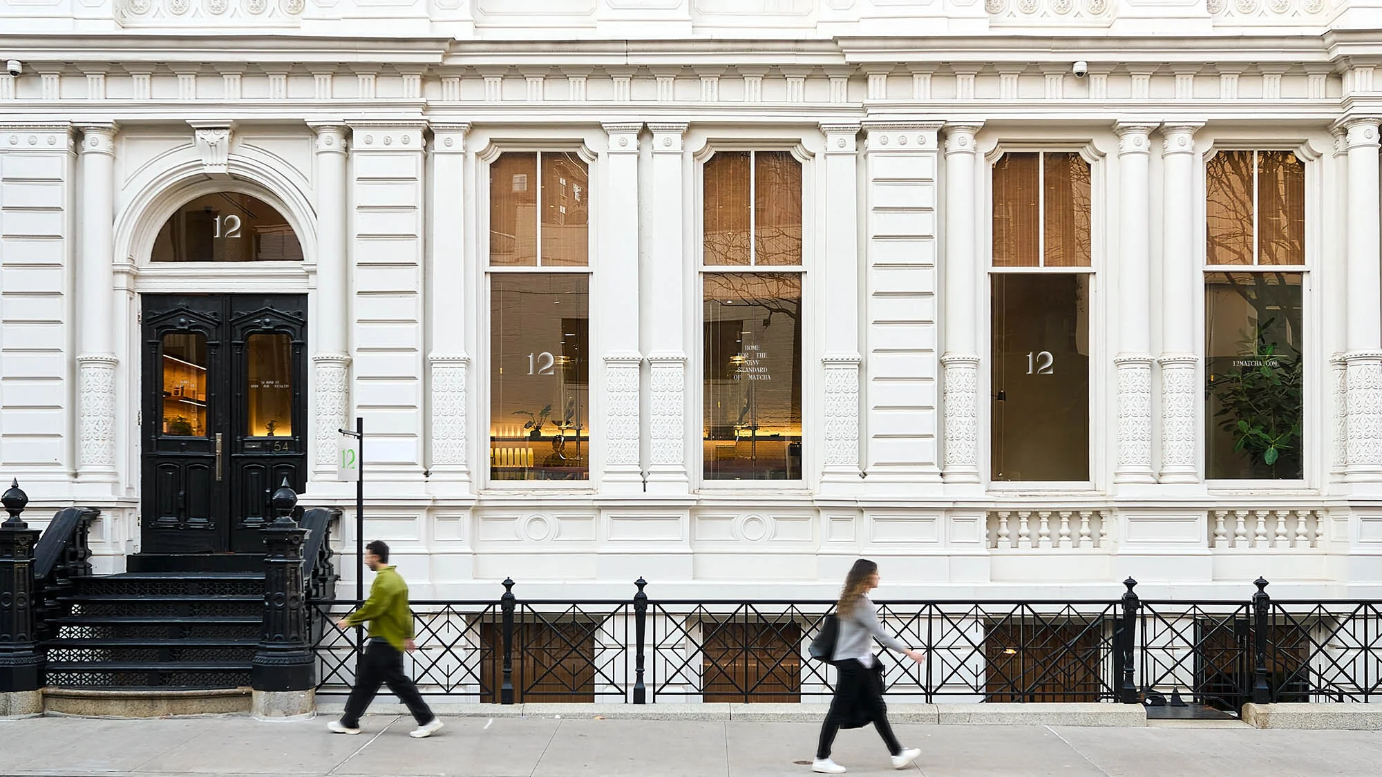 Exterior of a white building with large windows and a dark door, people walking on the sidewalk. Building is marked with the number 12.