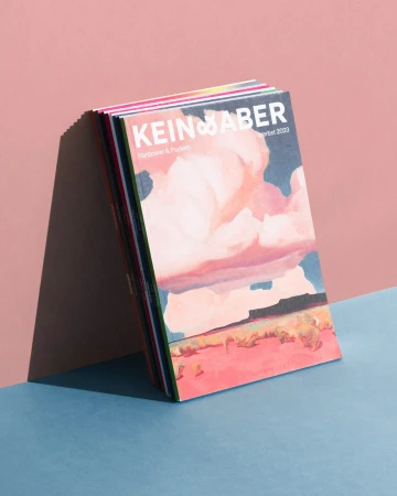 Stack of KEIN & ABER magazines featuring painted landscape covers against a pink background, displayed on a blue surface, showcasing color harmony.
